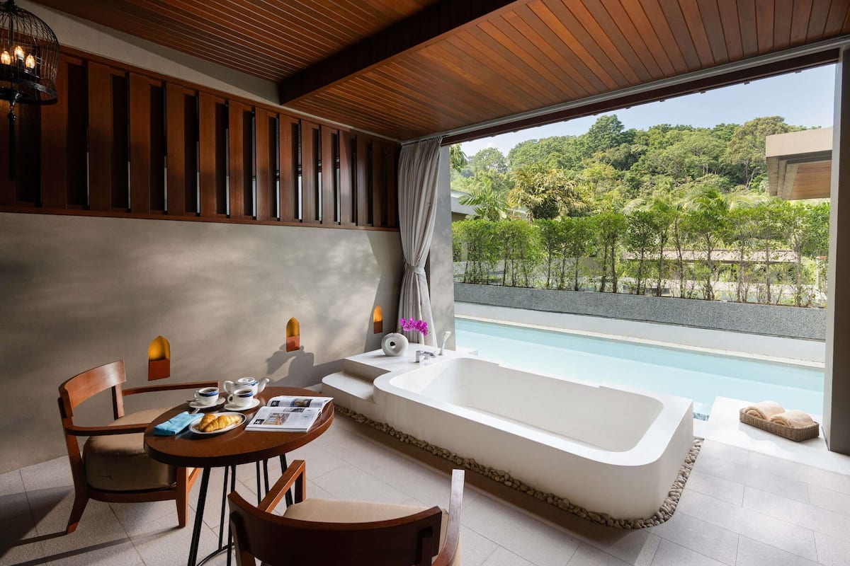 A modern bathroom with a large bathtub, a sink, and a table with chairs. There is a view of a pool and greenery outside.