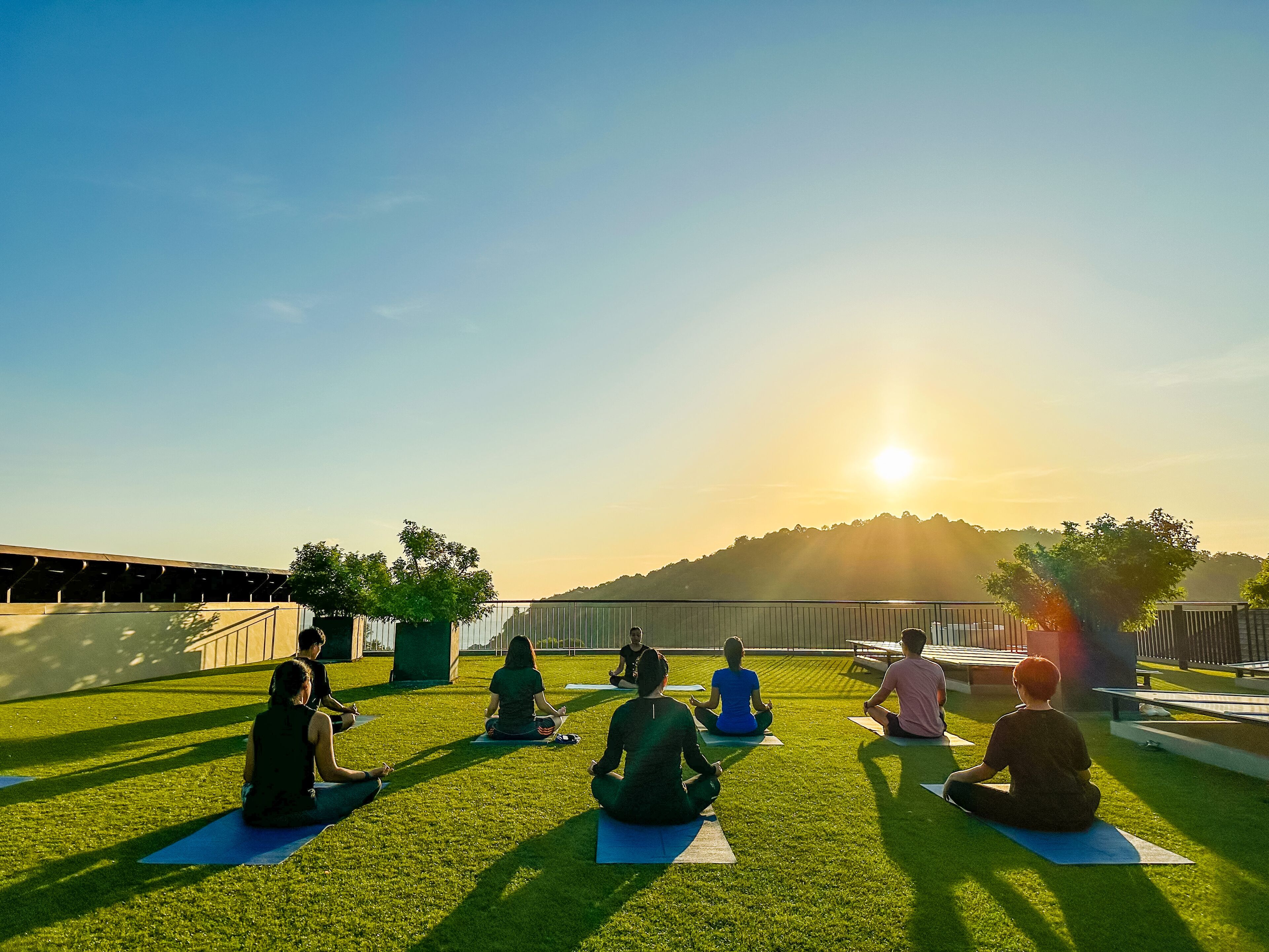 Een groep mensen mediteert op yogamatten in een openlucht omgeving, met een heldere lucht en een zonsondergang op de achtergrond.