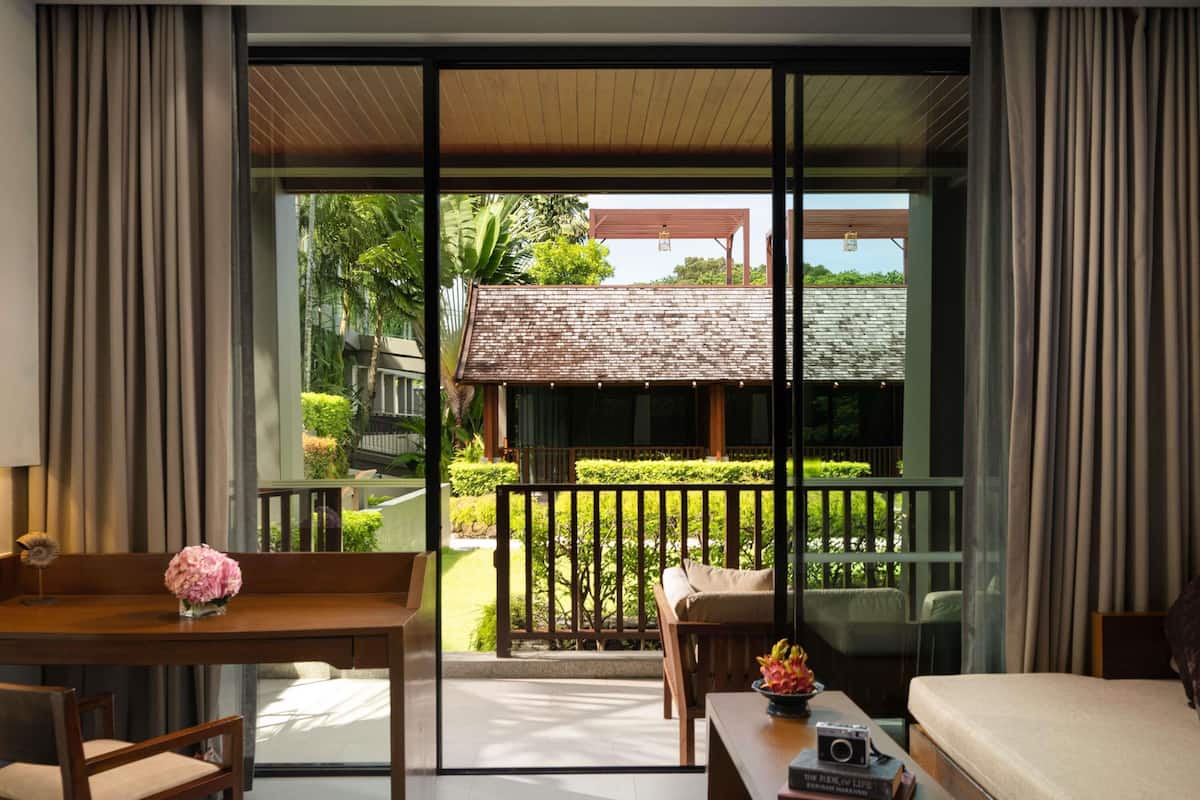 A modern room with a large sliding glass door leading to a balcony with a view of a garden and a building. There is a wooden table with chairs and a sofa.