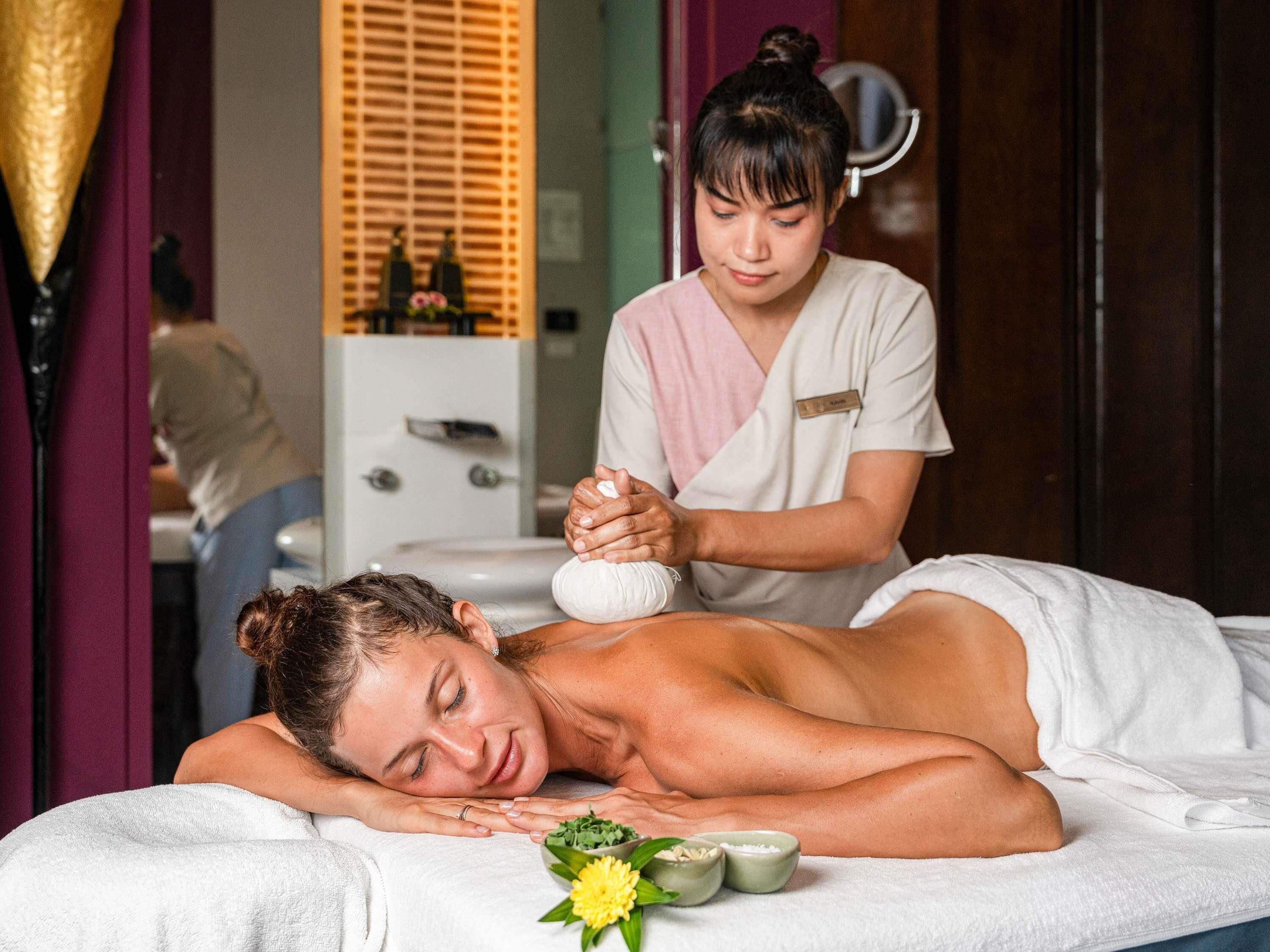 A massage therapy session in a room with a bed, a small table with a flower, and a cabinet in the background.