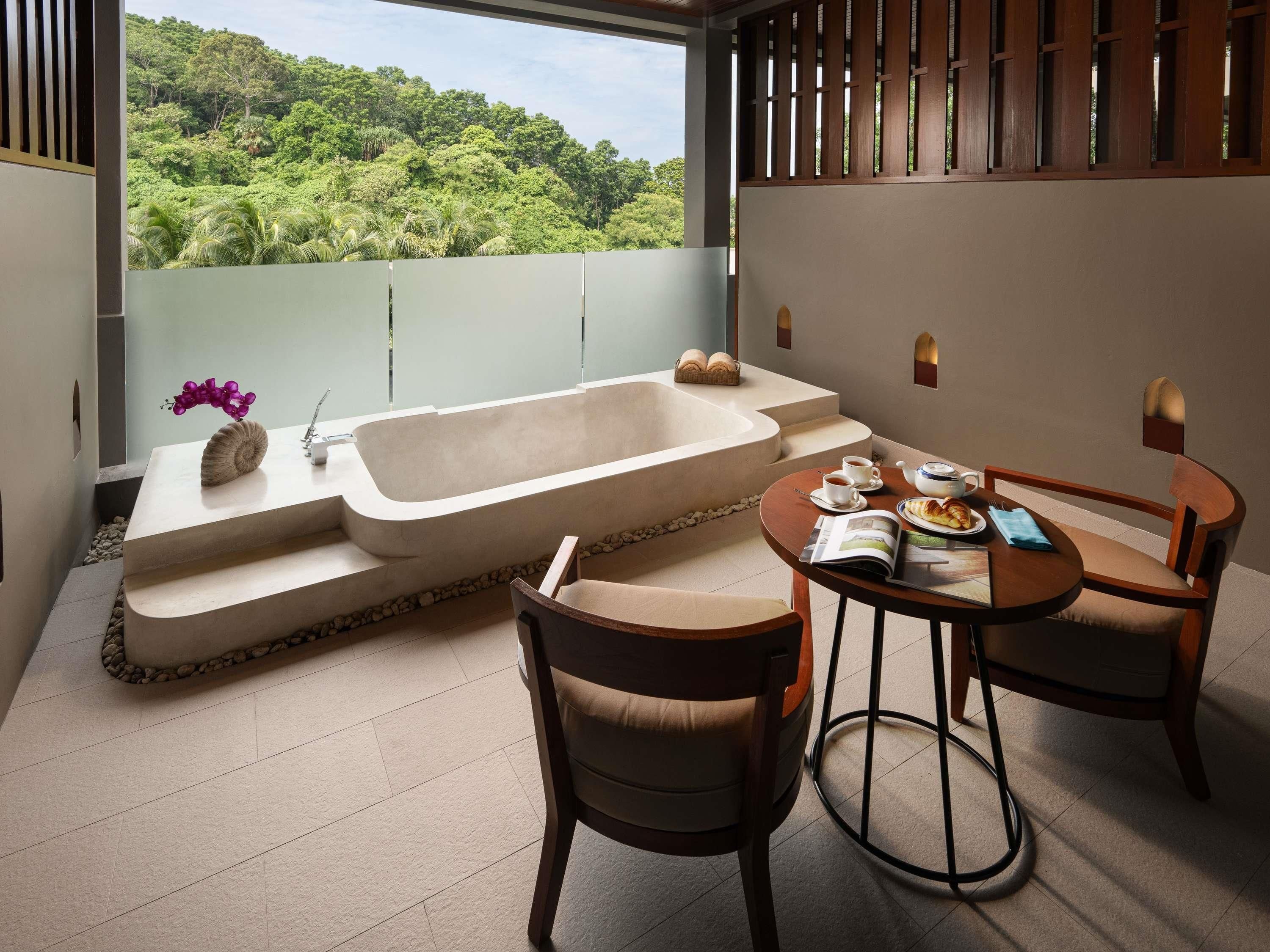 A modern bathroom with a bathtub, a sink, and a table with chairs. There is a view of a lush green landscape through a window.