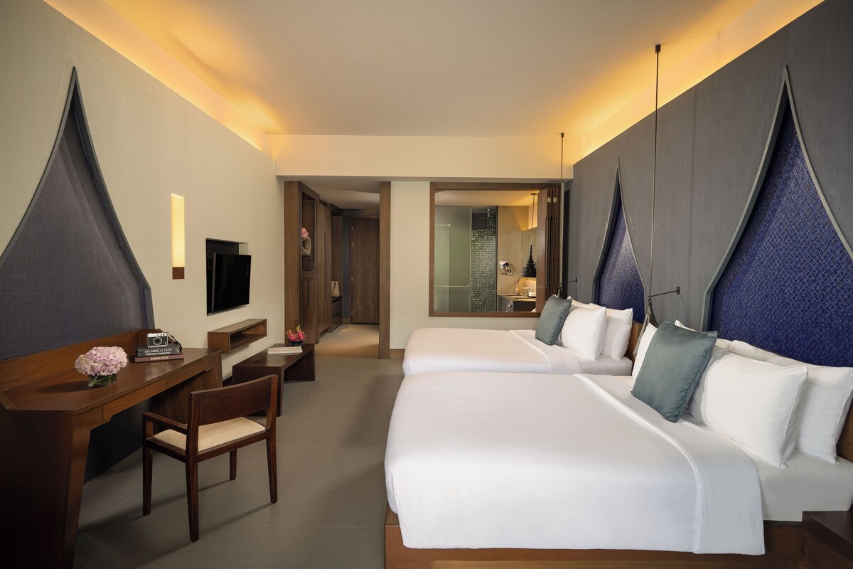 Habitación de hotel con dos camas, un escritorio, una silla, un televisor y un baño visible a través de una puerta abierta.