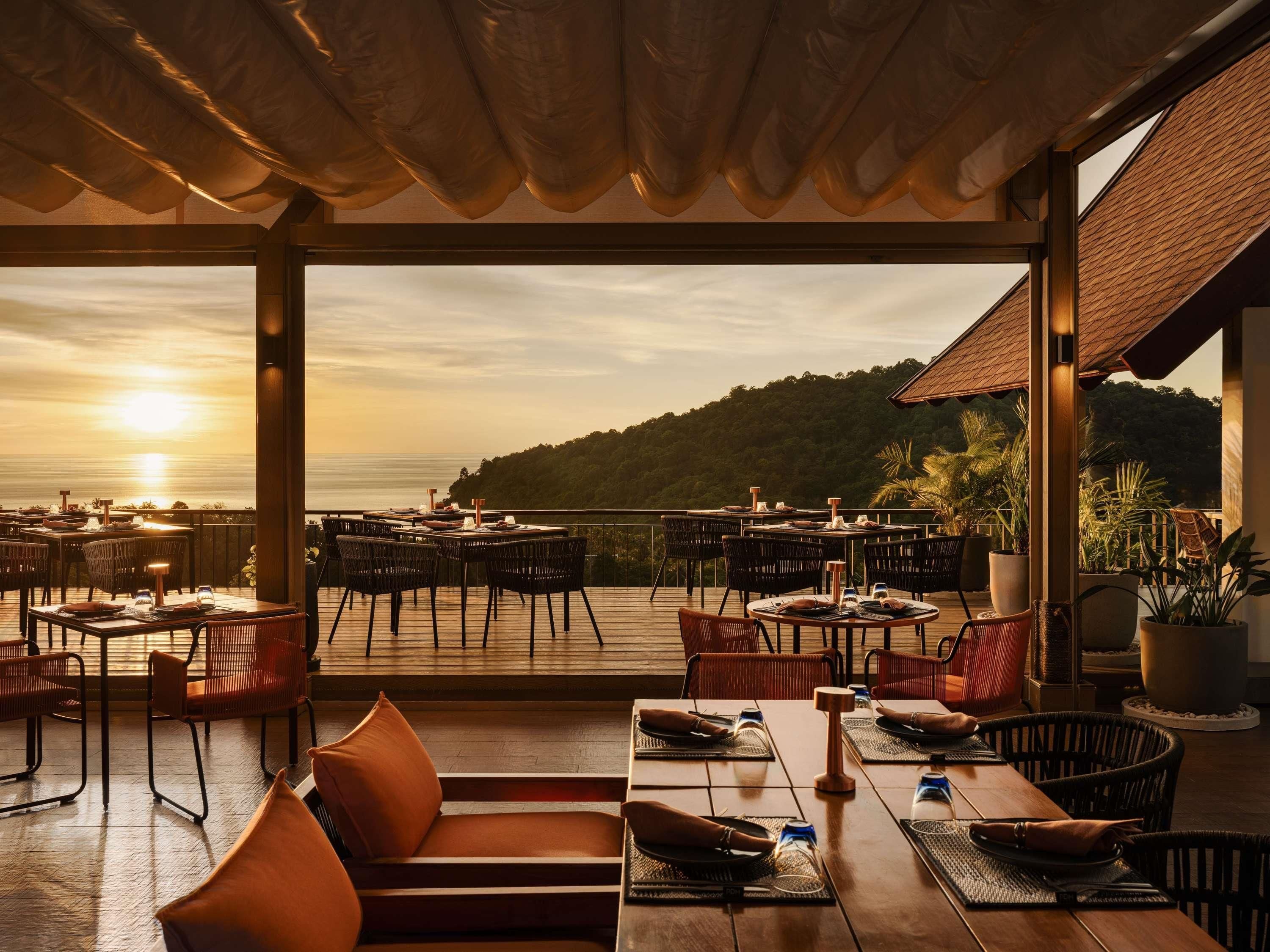Un espace de restauration en plein air, avec des tables et des chaises, offrant une vue panoramique sur l’océan et les collines.