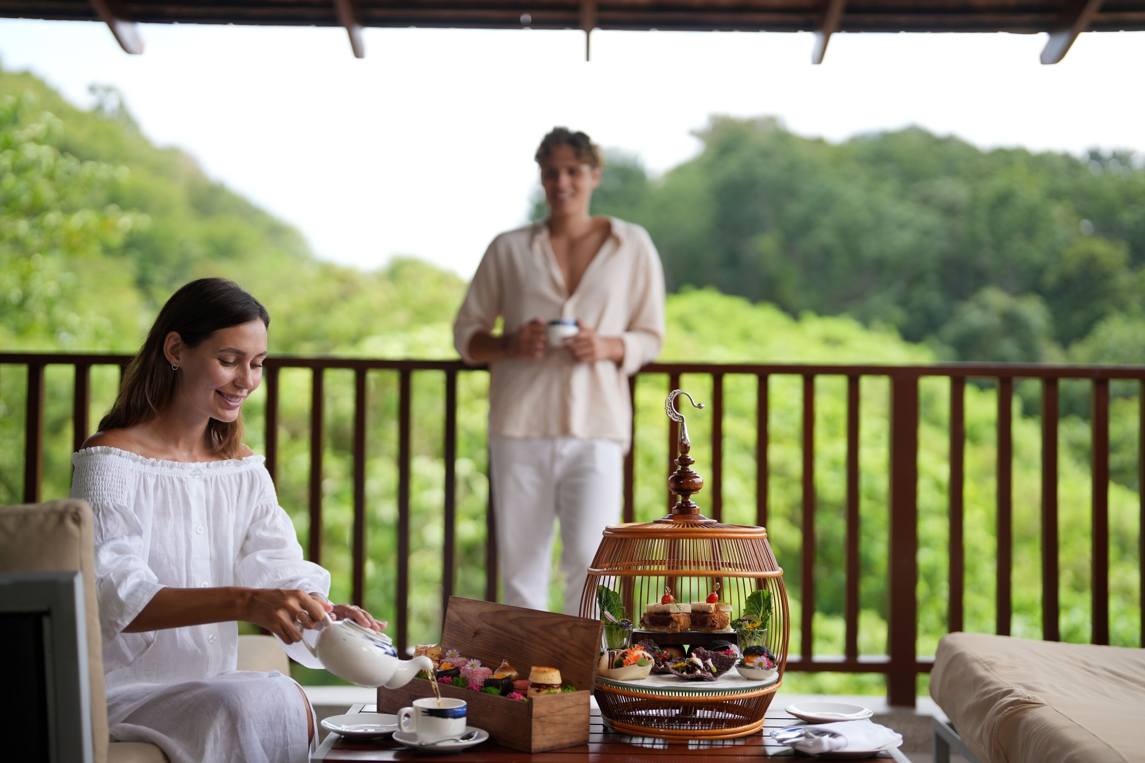 A woman pouring tea into a cup on a wooden table, with a tiered dessert display and a man in the background holding a cup.