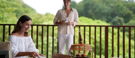 A woman pouring tea into a cup on a wooden table, with a tiered dessert display and a man in the background holding a cup.