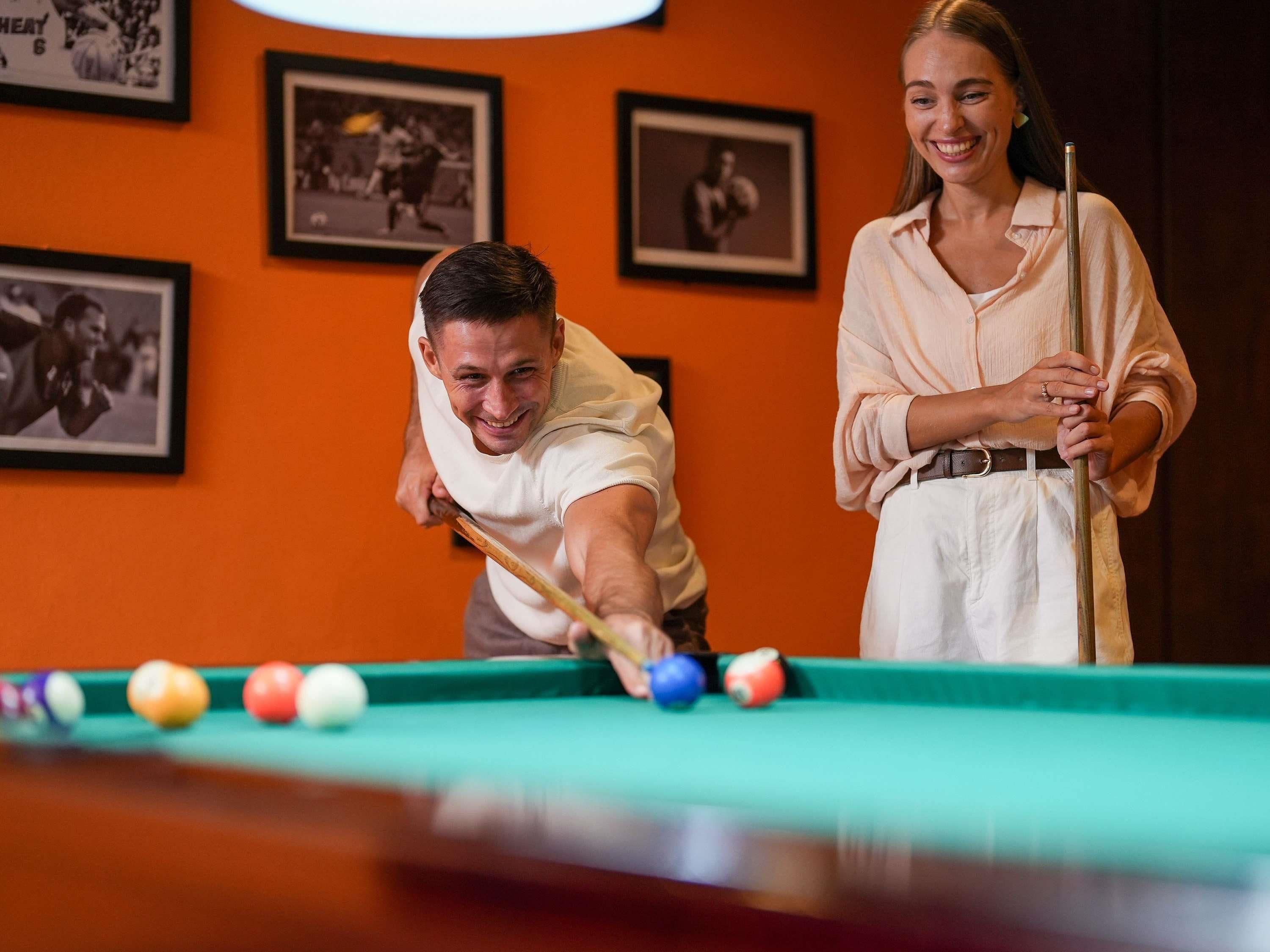 Two people playing pool in a room with orange walls and framed pictures.
