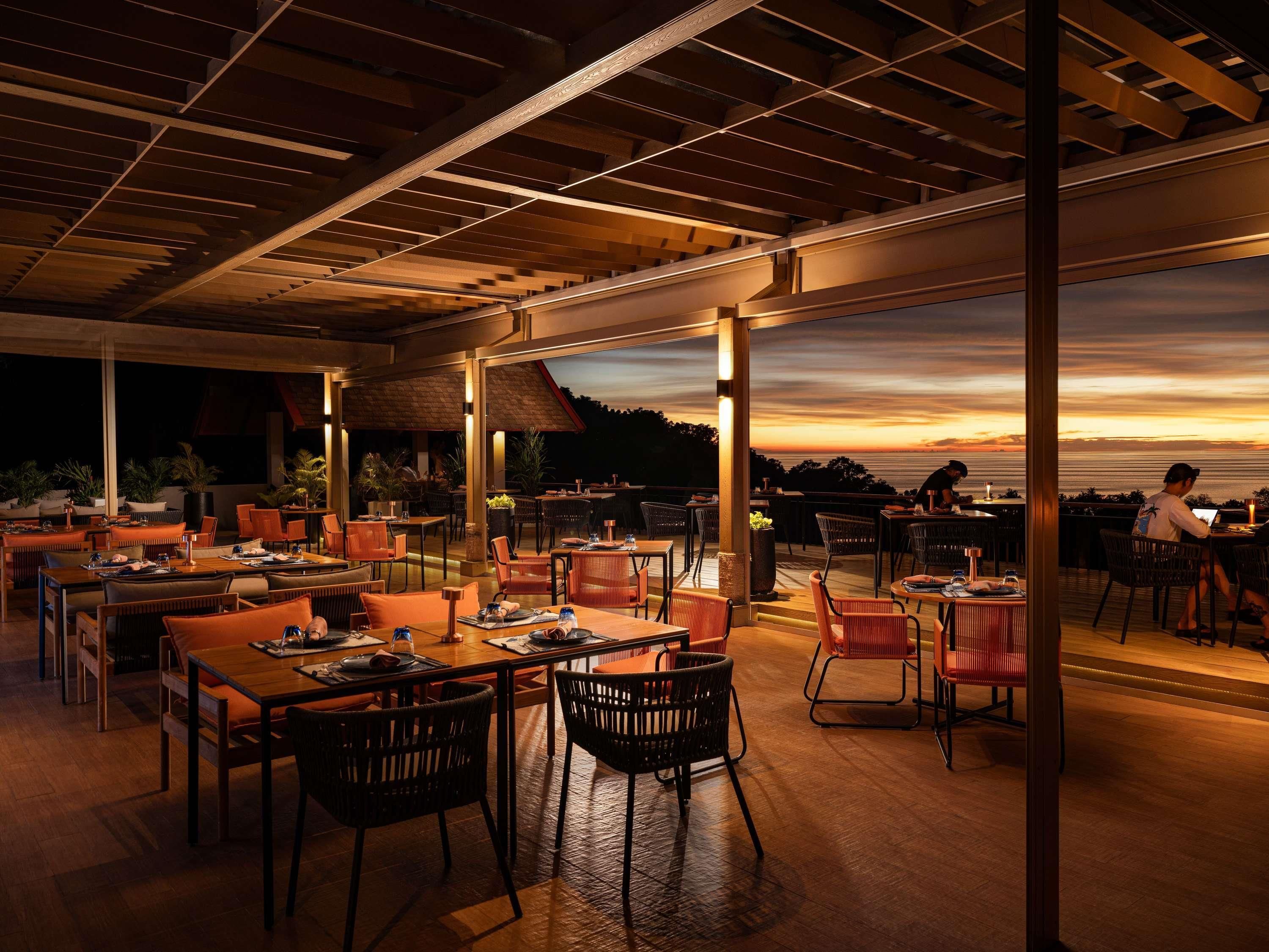An outdoor dining area with tables and chairs, a view of the sunset, and a wooden ceiling structure.