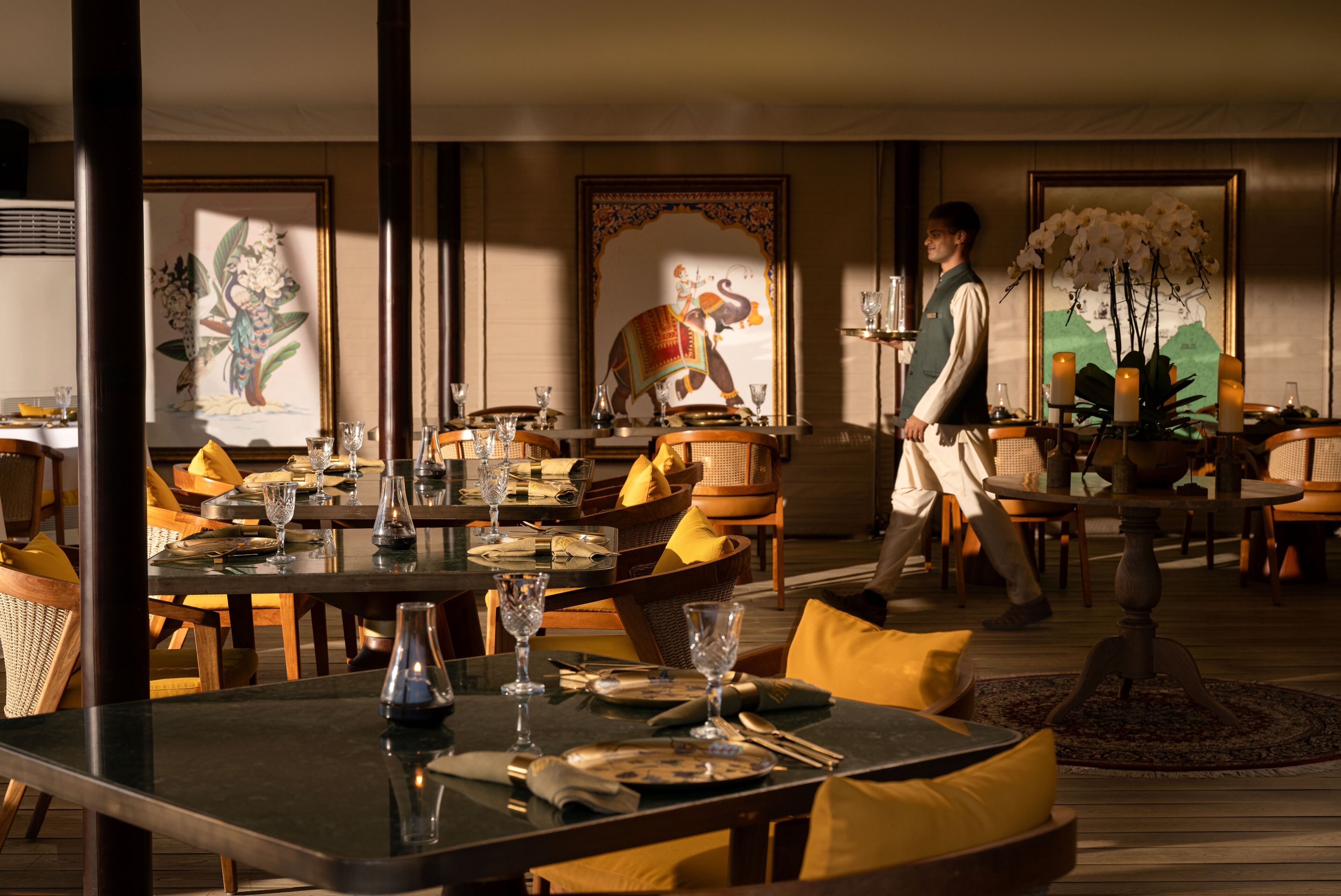 A well-lit dining area with tables set for a meal, featuring artwork on the walls and a waiter present.