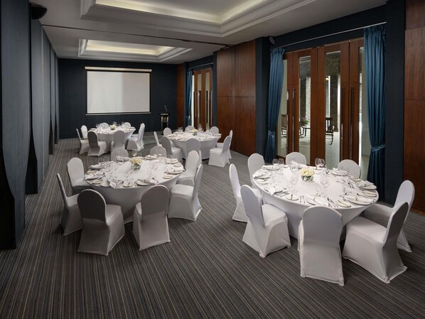 Un salón de conferencias con varias mesas redondas, sillas blancas y una pantalla para proyector.