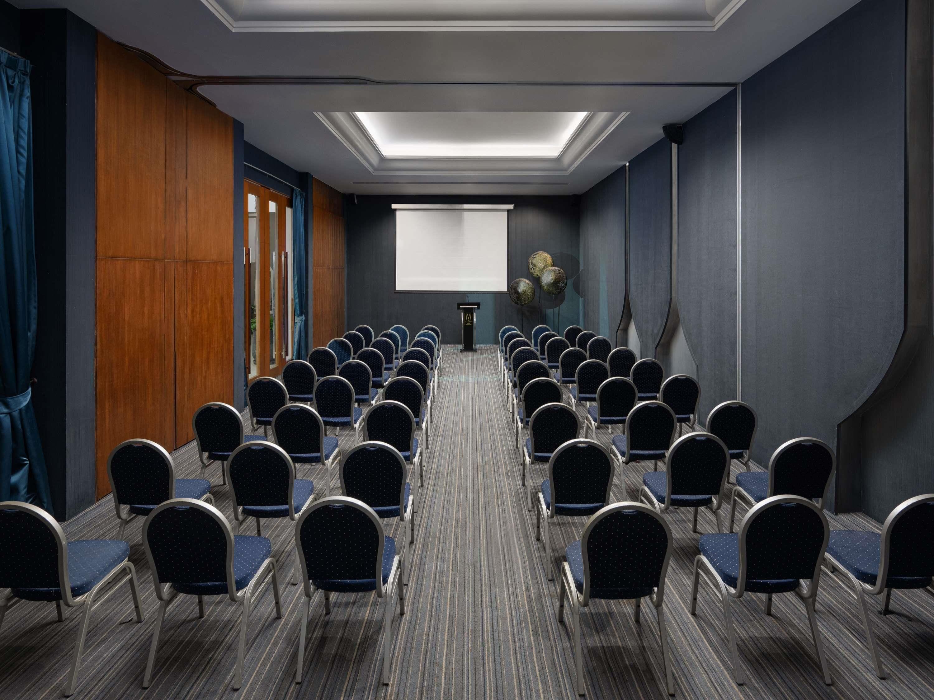 Un salón de conferencias con filas de sillas, una pantalla para proyector y un atril.
