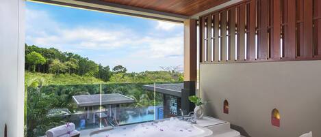 A modern bathroom with a large bathtub, a seating area with a chair and table, and a view of lush greenery outside.