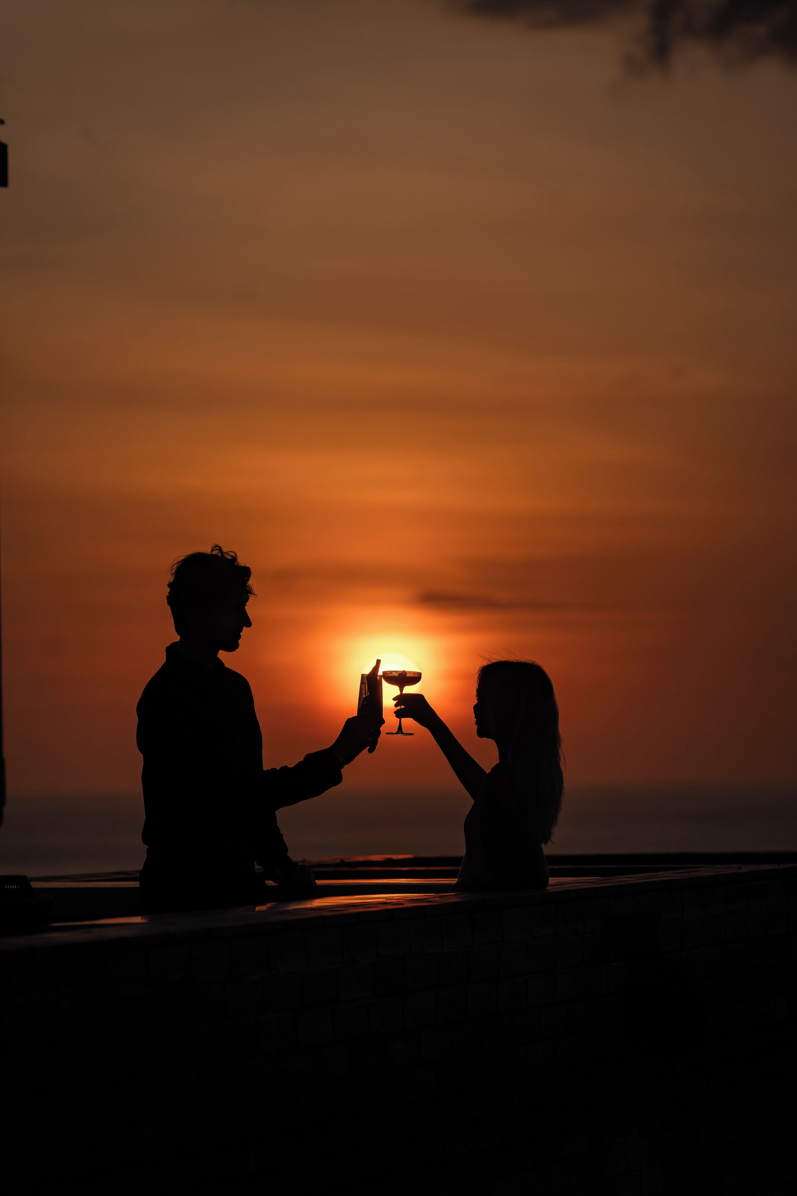 Silhuetter af to personer, der skåler med et glas mod en solnedgang.