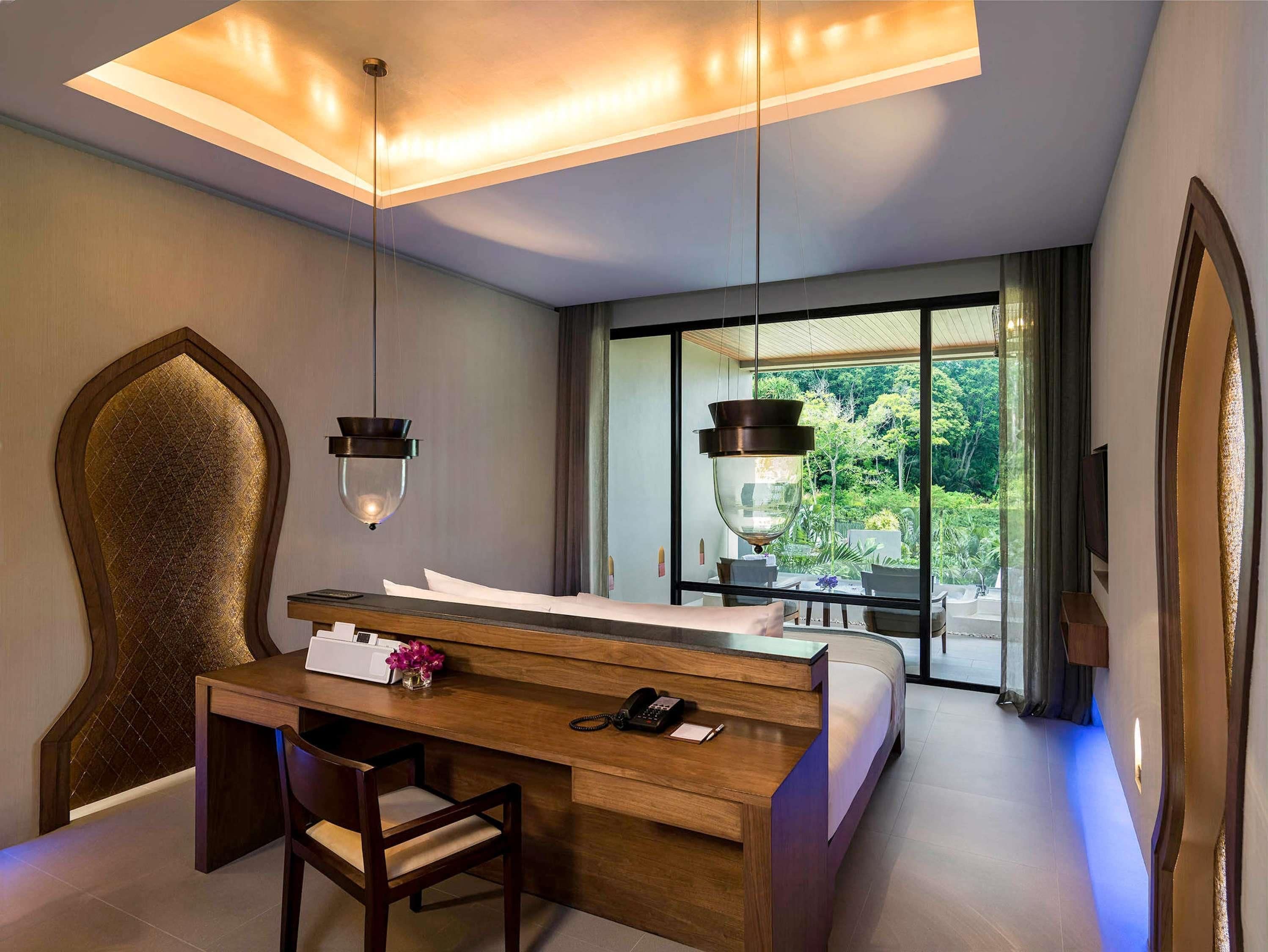 Une chambre d’hôtel moderne équipée d’un lit, d’un bureau et d’une chaise, d’une grande fenêtre donnant sur un paysage verdoyant et d’un luminaire suspendu.