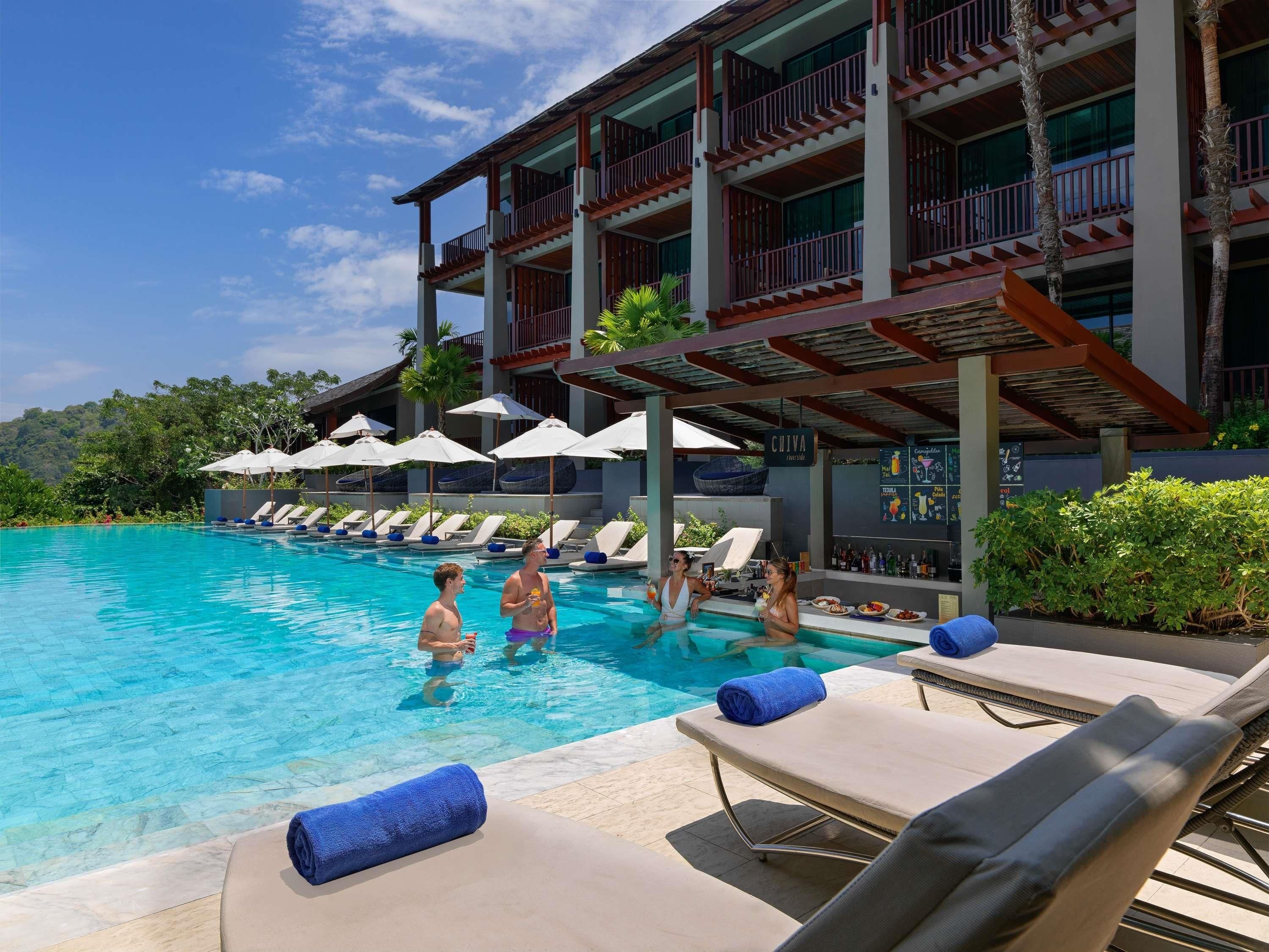 A pool area with lounge chairs and umbrellas, a bar, and a building with multiple balconies.