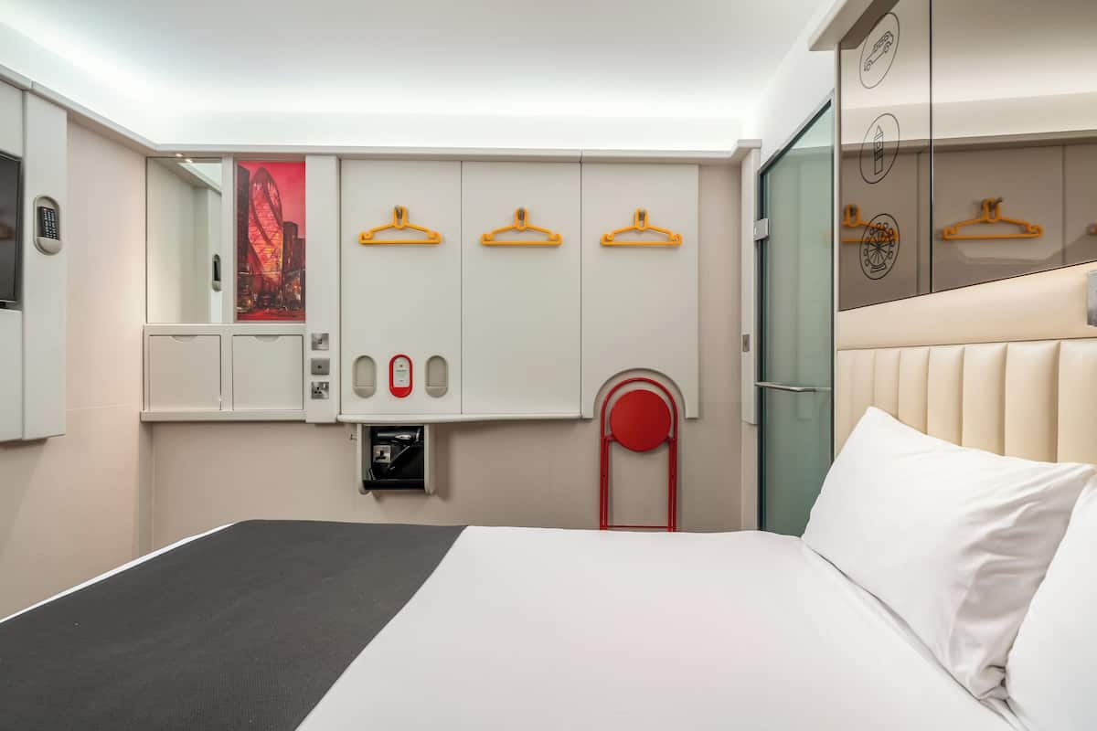 A hotel room with a bed, a wardrobe with hangers, a mirror, and a red chair.