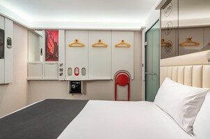 Una habitación de hotel con cama, armario con perchas, espejo y una silla roja.