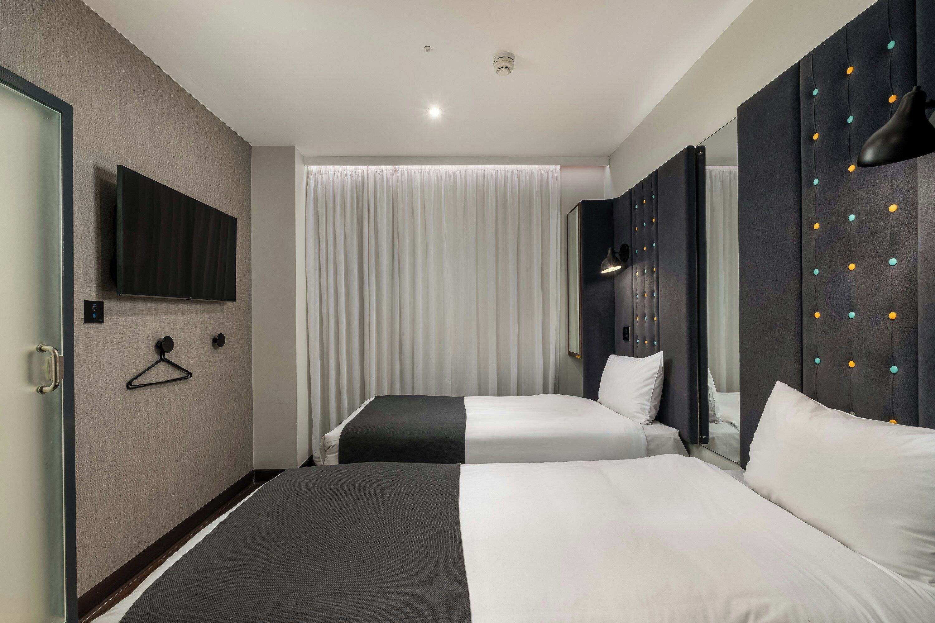 Habitación de hotel con dos camas, un televisor de pantalla plana, un espejo con luces decorativas y una ventana con cortinas.