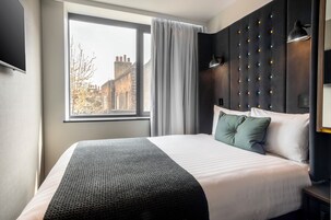 Ein Hotelzimmer mit einem großen Bett, einem Fenster mit Vorhängen, Blick auf die Stadt, einem gepolsterten Kopfteil und einer Nachttischlampe.