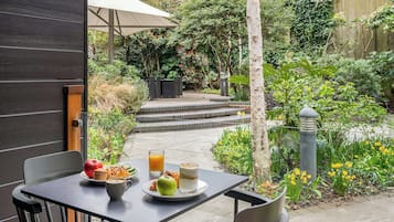 Un patio con una mesa dispuesta para el desayuno, que incluye un plato con una manzana, un muffin y un croissant, un vaso de jugo de naranja y un vaso de leche con cereal.