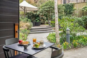 A patio with a table set for breakfast, including a plate with an apple, muffin, and croissant, a glass of orange juice, and a glass of milk with cereal.
