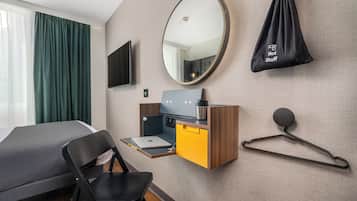 A hotel room with a bed, a desk with a laptop, a mirror, a TV, and a hanging bag labeled "Hot Stuff".