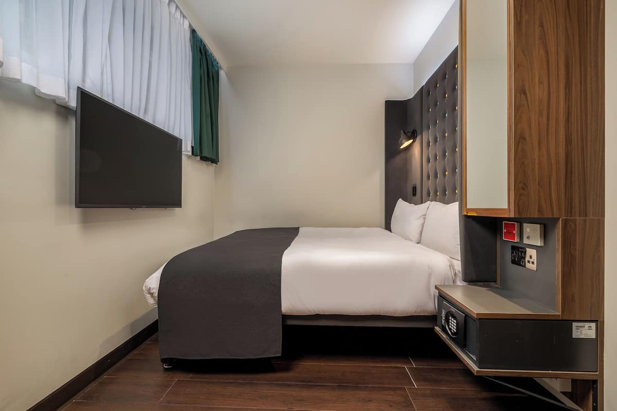 A hotel room with a bed, a flat-screen TV mounted on the wall, a wooden headboard, and a small bedside table.