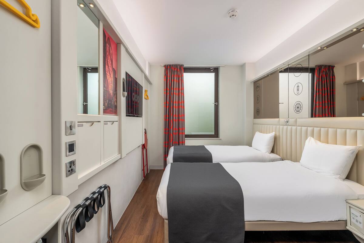 A hotel room with two beds, a large mirror, a window with red curtains, and a radiator.