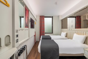 A hotel room with two beds, a large mirror, a window with red curtains, and a radiator.