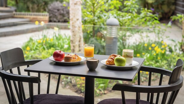 Ein Tisch mit Frühstücksangeboten, darunter ein Muffin, ein Croissant, ein Apfel und ein Glas Orangensaft, auf einem Balkon mit Blick auf einen Garten.