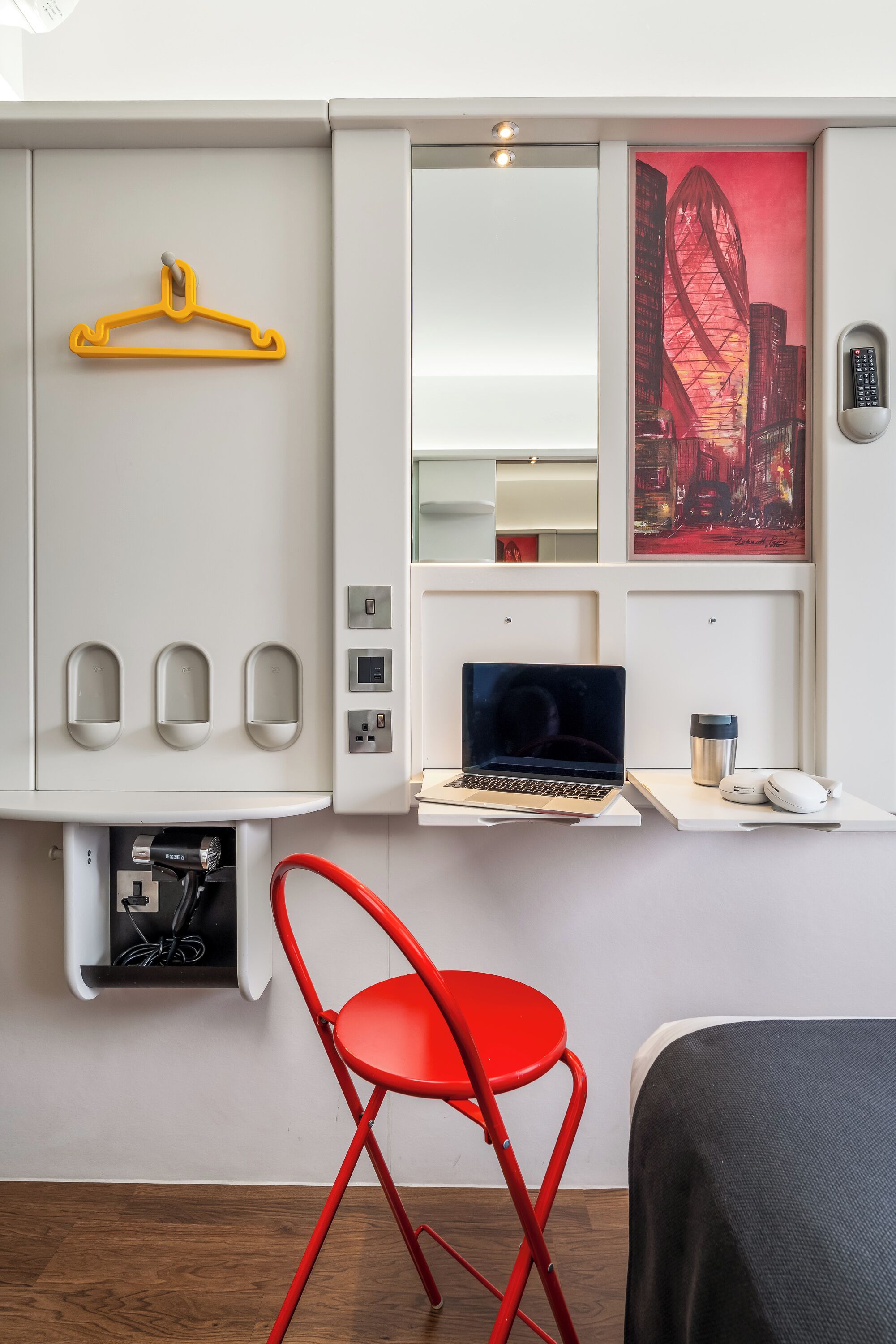 Habitación de hotel con escritorio, computadora portátil, silla roja y un cuadro de un paisaje urbano.