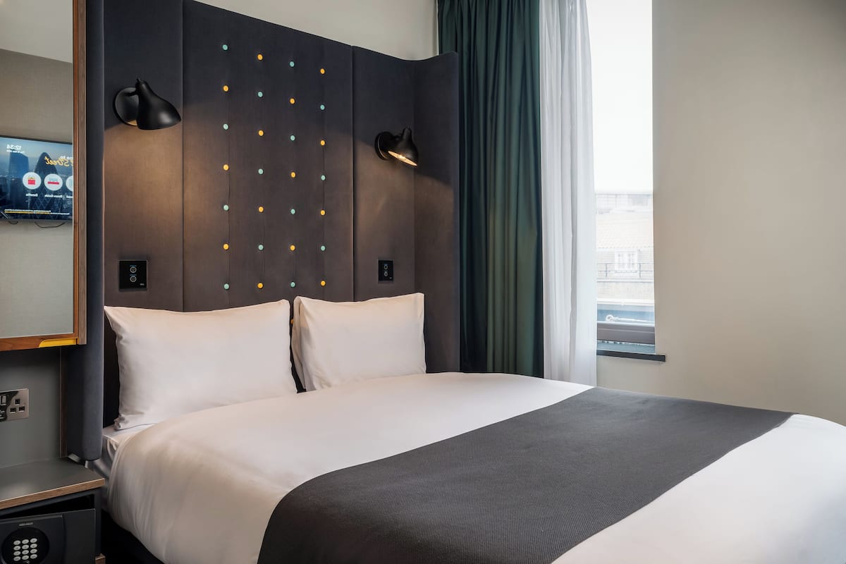 A hotel room with a bed, a TV, a control panel, and a window with curtains.