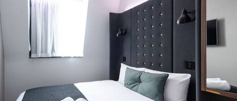 A modern hotel room with a neatly made bed, a large window with curtains, a wall-mounted TV, and a bedside lamp.