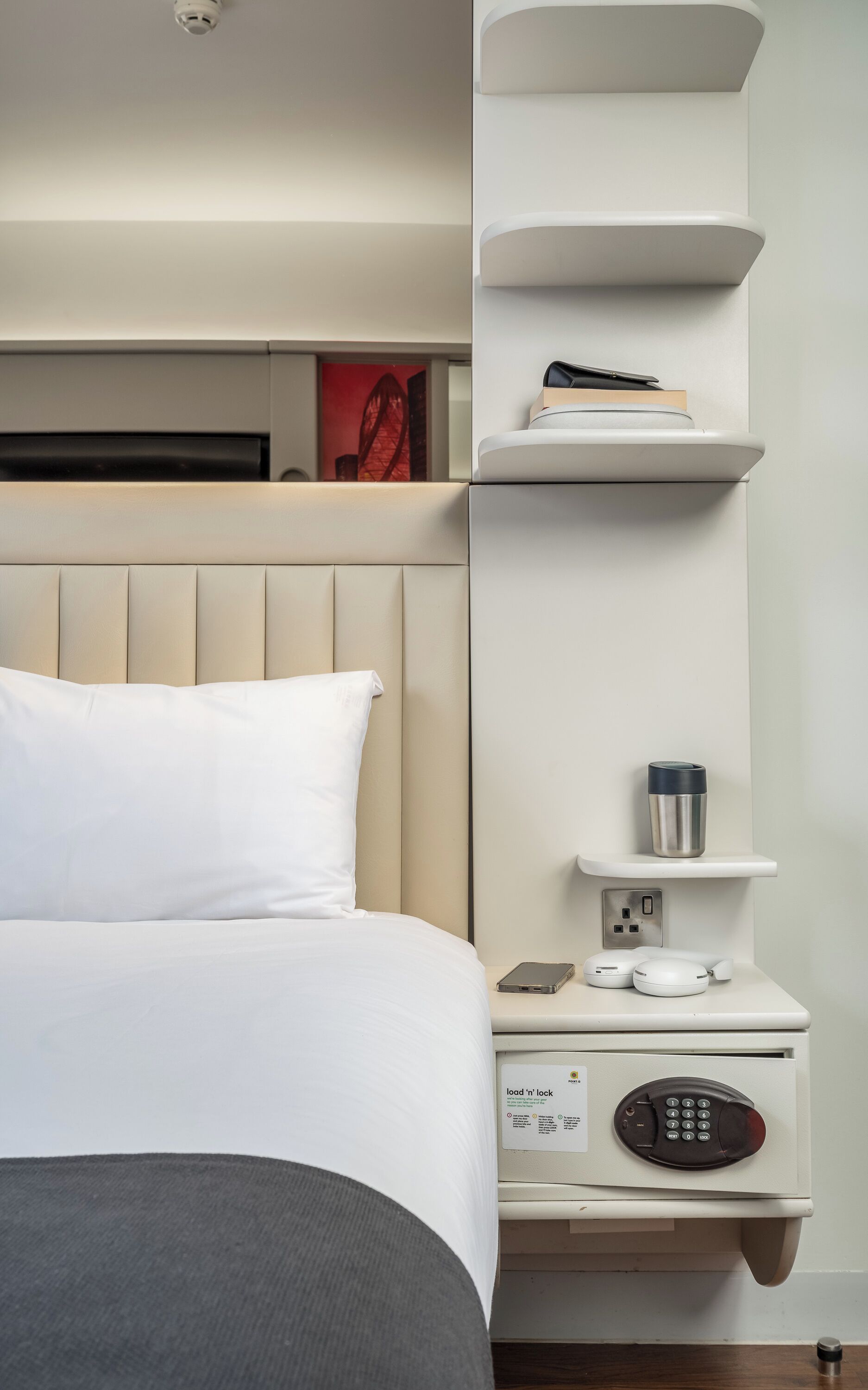 A hotel room with a bed, a bedside table with a phone, a coffee maker, and a shelf with books and a laptop.