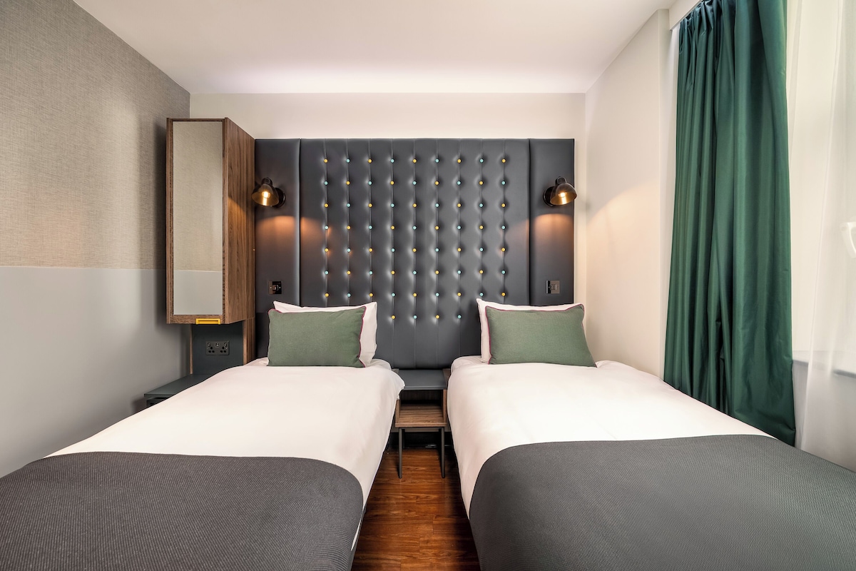 A hotel room with two beds, a wooden headboard, bedside lamps, and a green curtain.