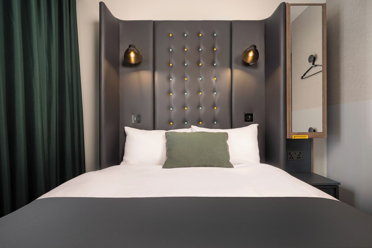 A modern hotel room with a large bed, a headboard with vertical light fixtures, a bedside table, and a mirror.