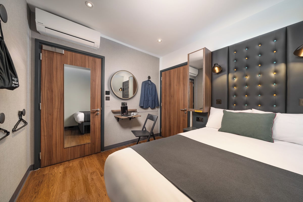 Ein modernes Hotelzimmer mit einem großen Bett, einem Holzschrank, einem Spiegel, einem kleinen Schreibtisch und einem Kleiderhaken.