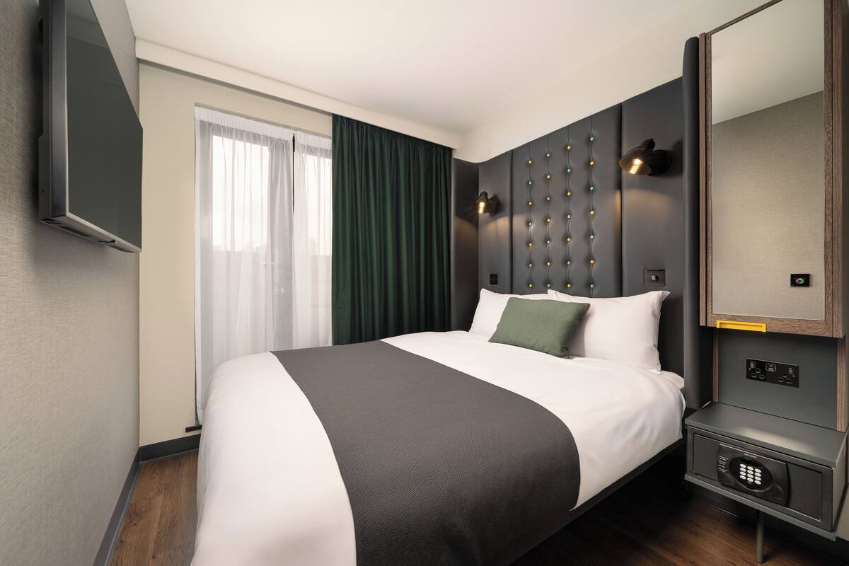 Ein modernes Hotelzimmer mit einem großen Bett, einem flachbildfernseher an der Wand, einem Nachttisch mit Lampe und einem fest eingebauten Safe.