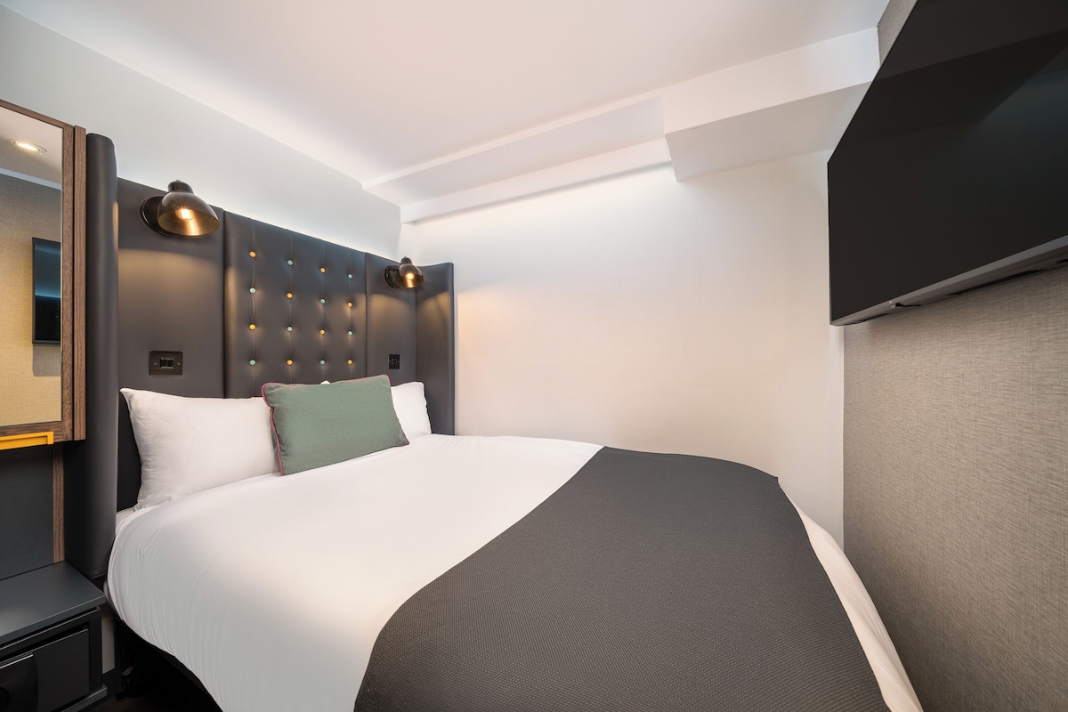A modern hotel room with a large bed, a headboard with a light fixture, a flat-screen TV, and a bedside table.