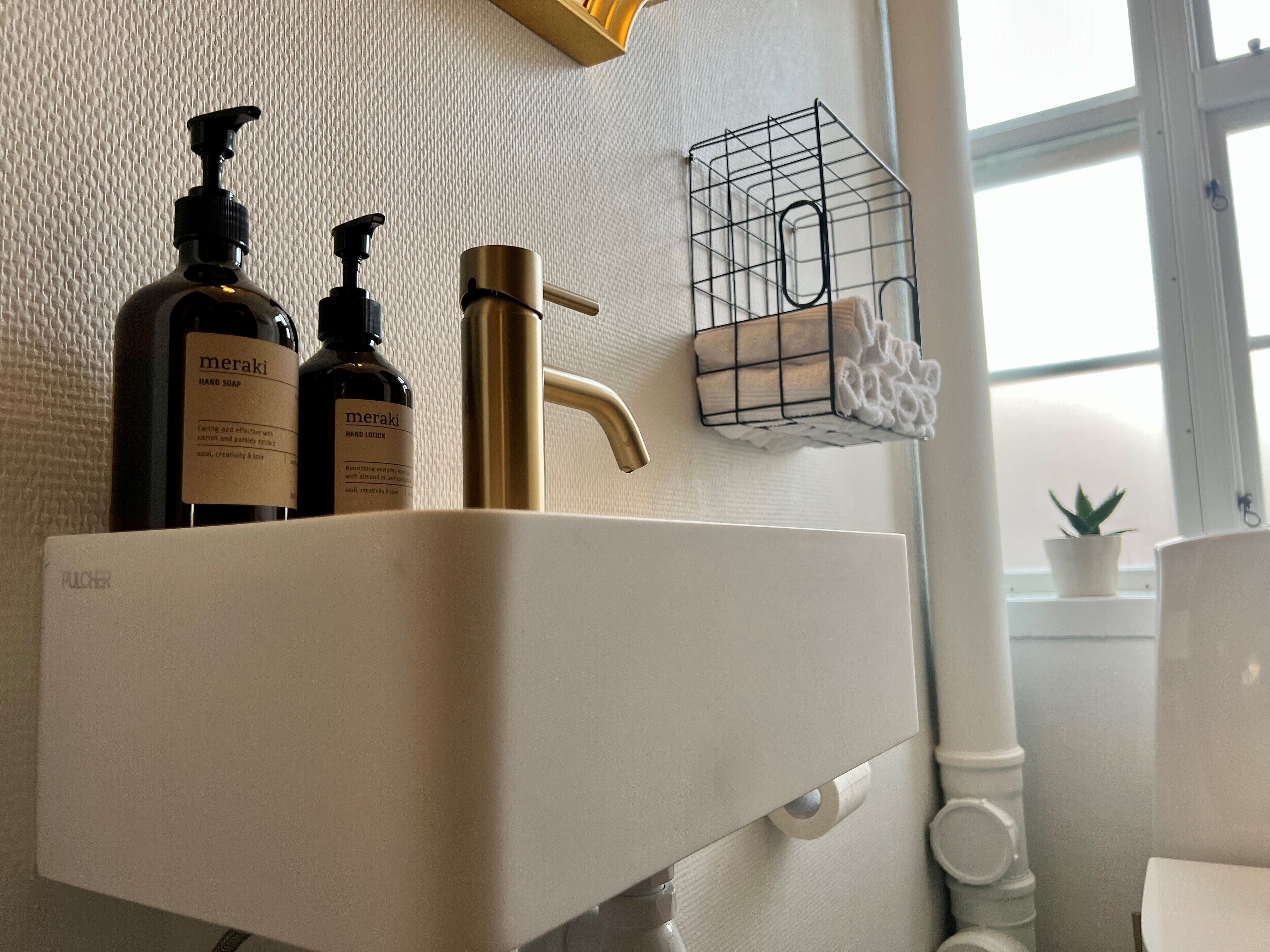 Een wastafel met twee flesjes Meraki handzeep en lotion, een gouden kraan en een handdoekhouder met een opgevouwen handdoek.