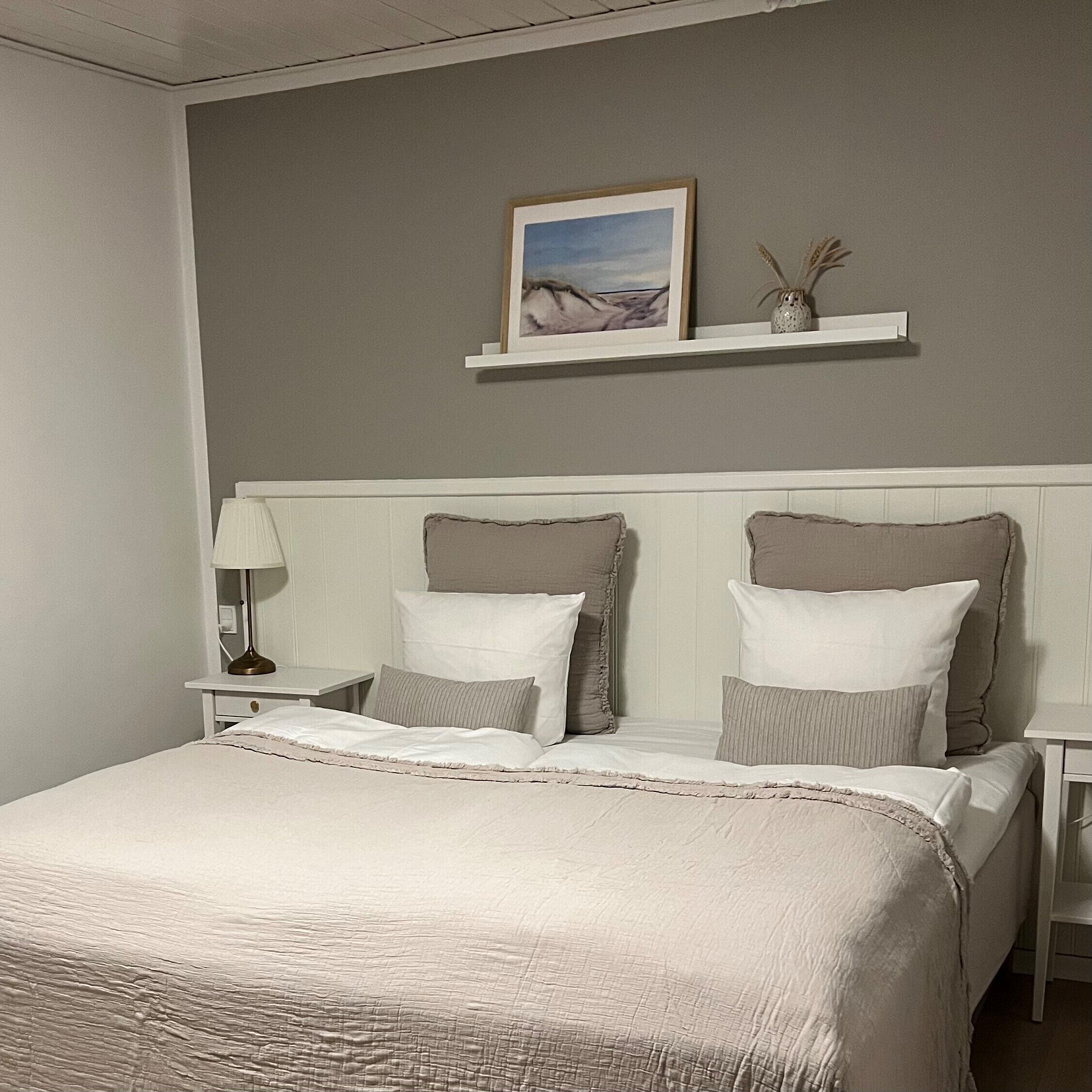 Una cama bien tendida con una paleta de colores blanco y beige, una mesita de noche con una lámpara, y una estantería con un cuadro enmarcado y un pequeño jarrón.