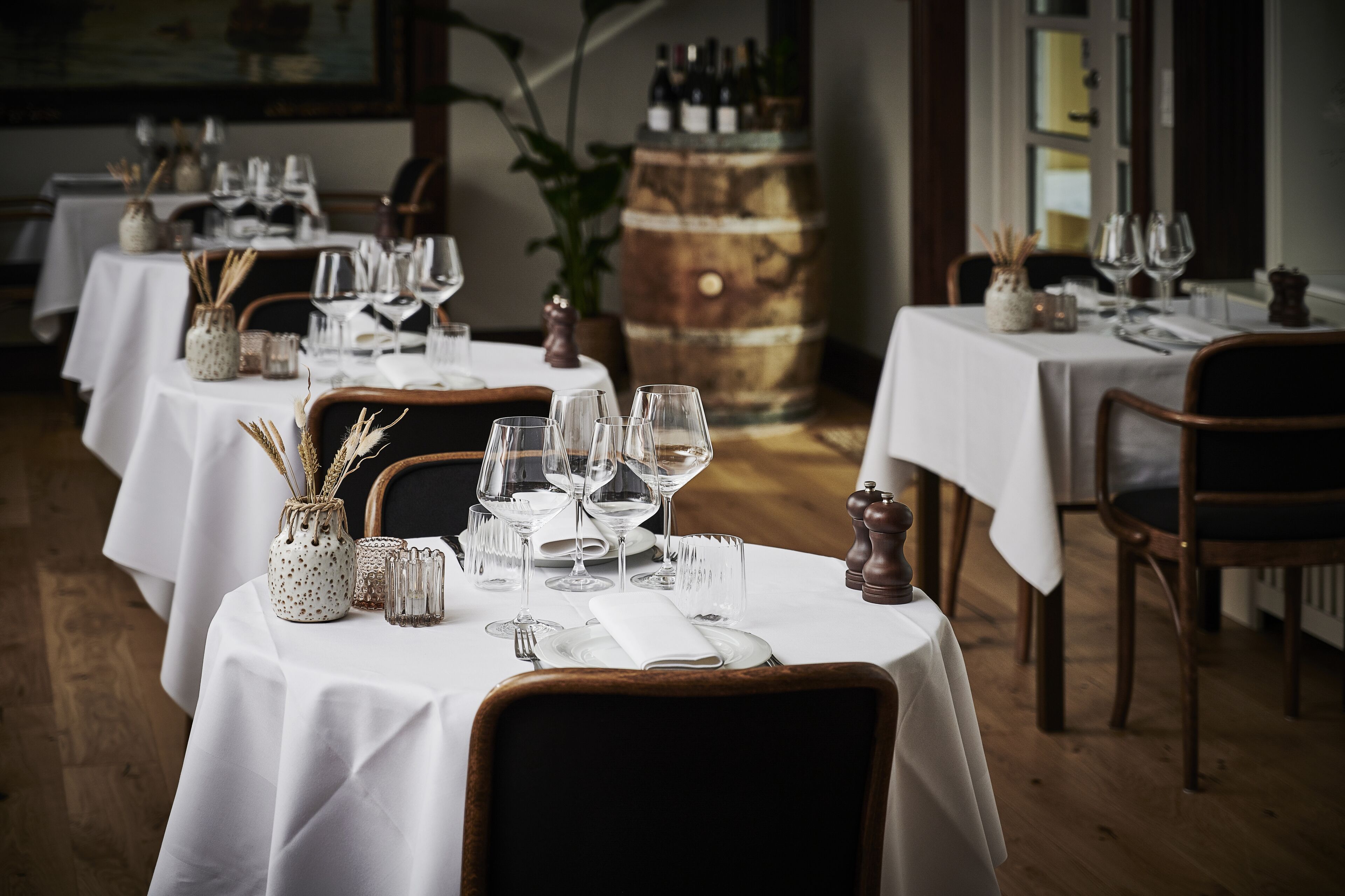 Un restaurante con varias mesas dispuestas para comer, con manteles blancos, copas de vino y cubiertos.