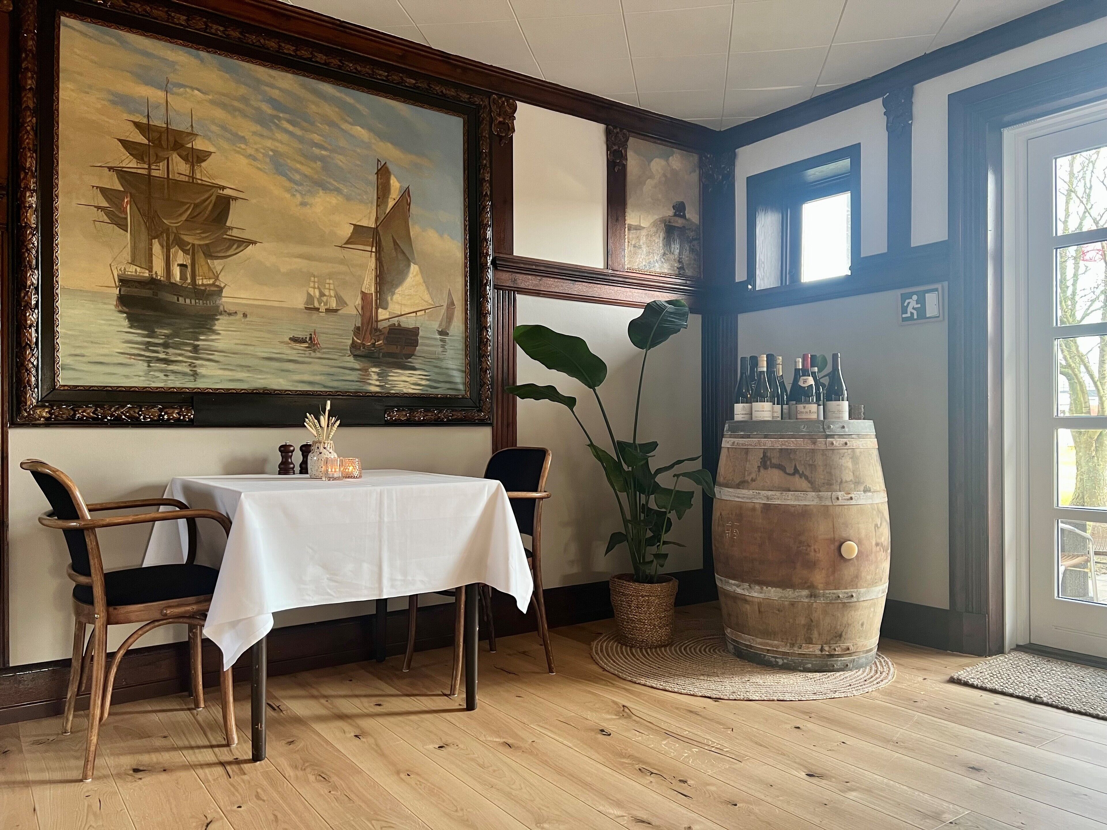 Un comedor con una mesa preparada para dos personas, un cuadro marino grande, un barril de madera y una planta en maceta.