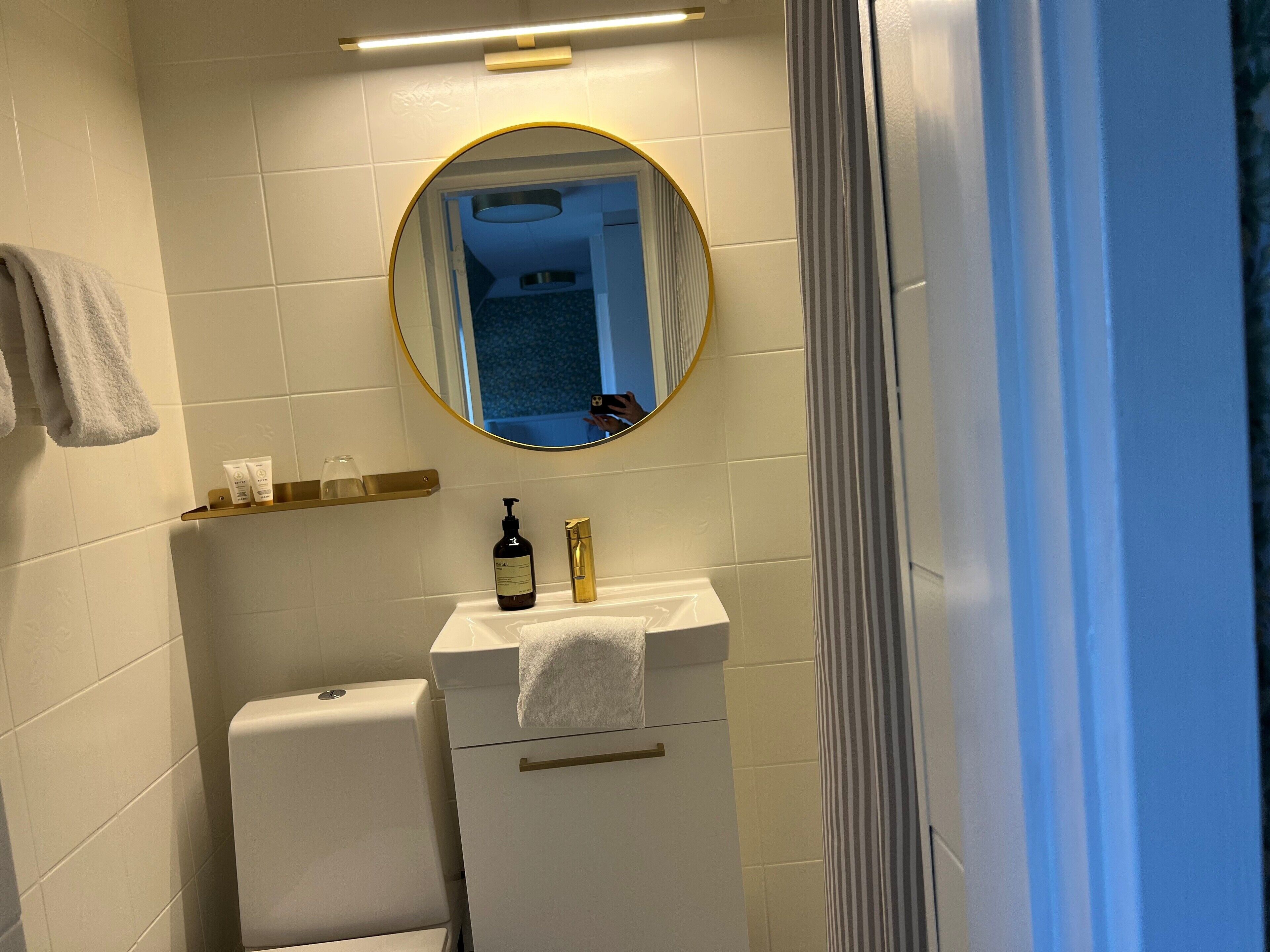 A bathroom with a white sink, a mirror, and a toilet. There is a towel hanging on the wall and a shelf with toiletries.