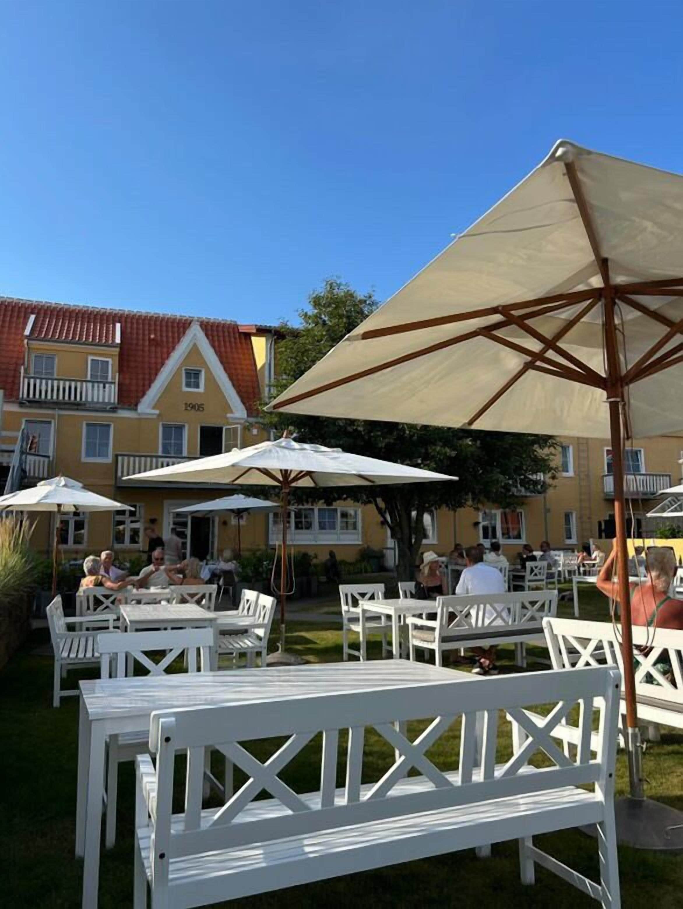 An outdoor seating area with white tables and chairs, shaded by large umbrellas, in front of a yellow building with a red roof and the number 1905.