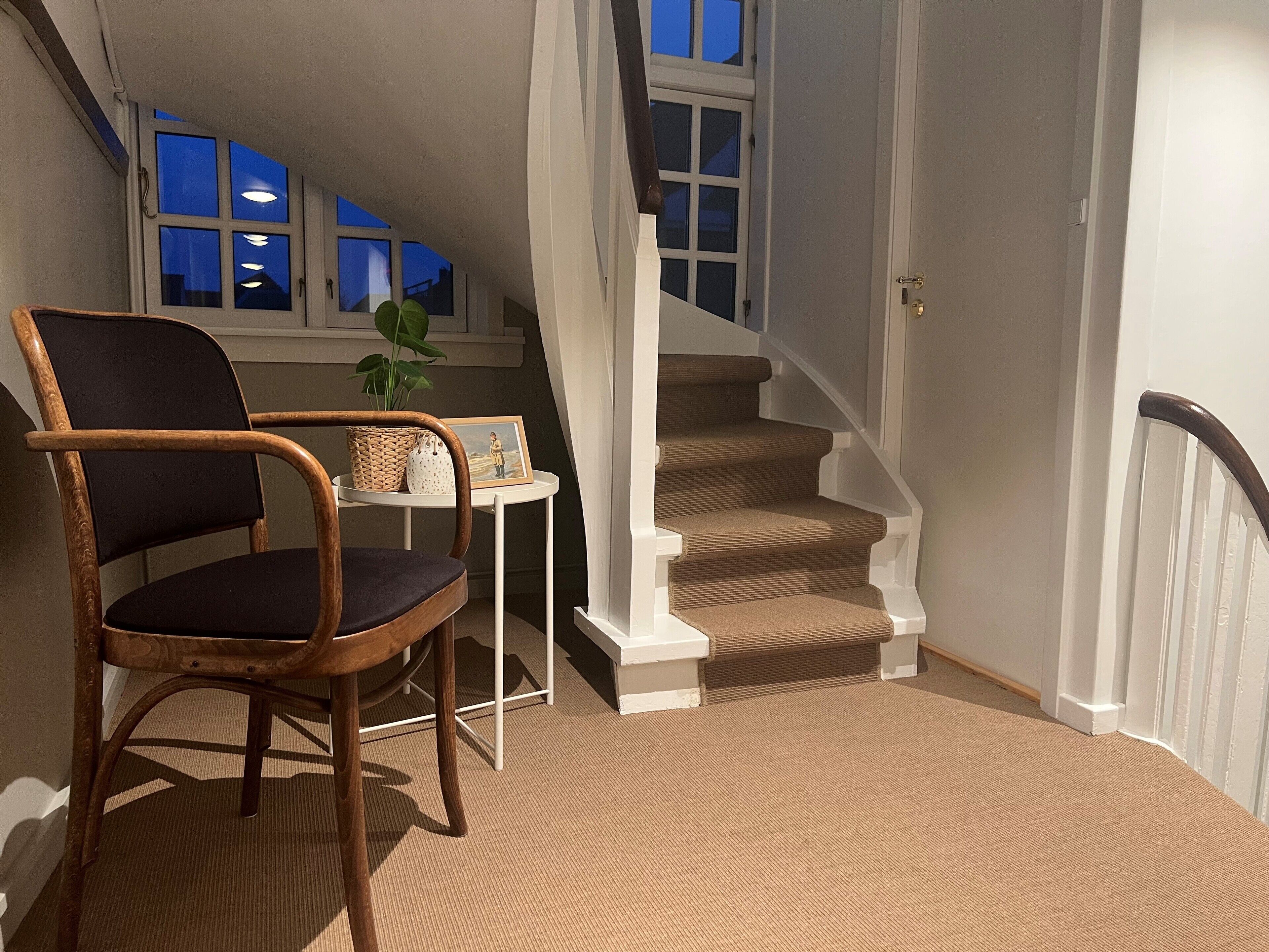 A cozy room with a wooden chair, a small table with a plant, and a staircase with beige carpet.