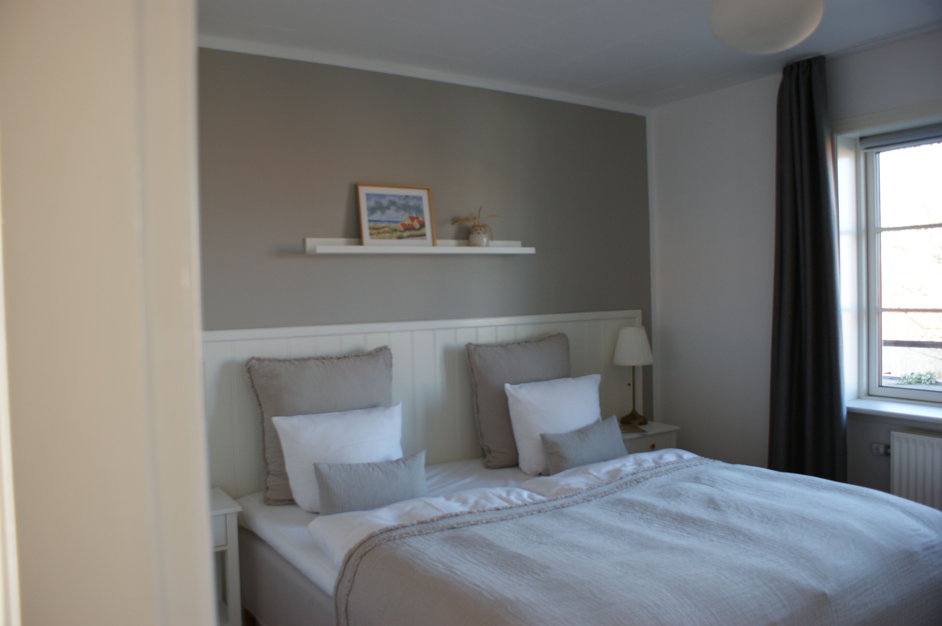 A bedroom with a bed, bedside tables, a nightstand, a wall-mounted shelf with a picture and a small plant, and a window with curtains.