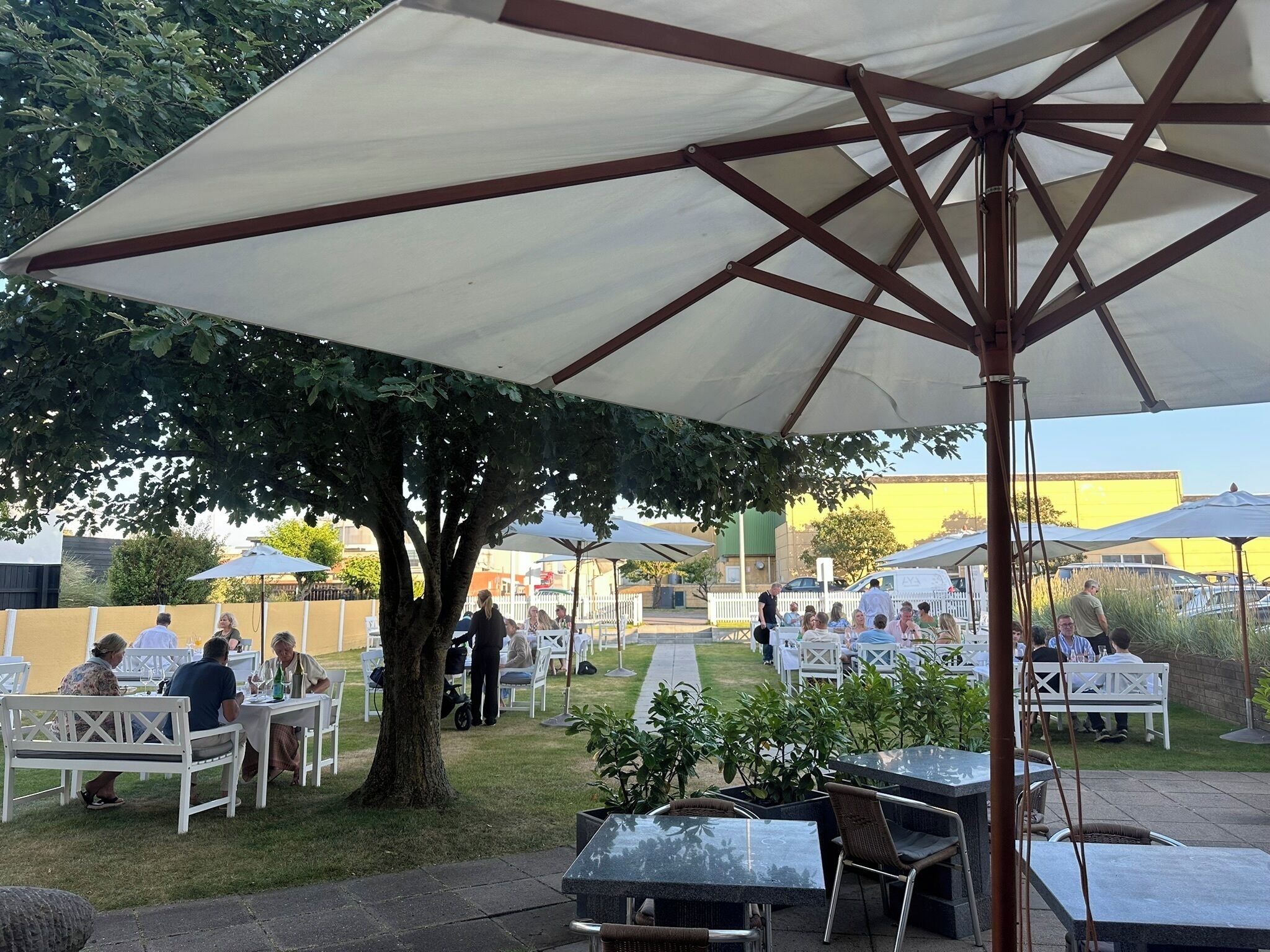 Un espacio exterior para comer con mesas y sillas bajo sombrillas, rodeado de follaje y un árbol.