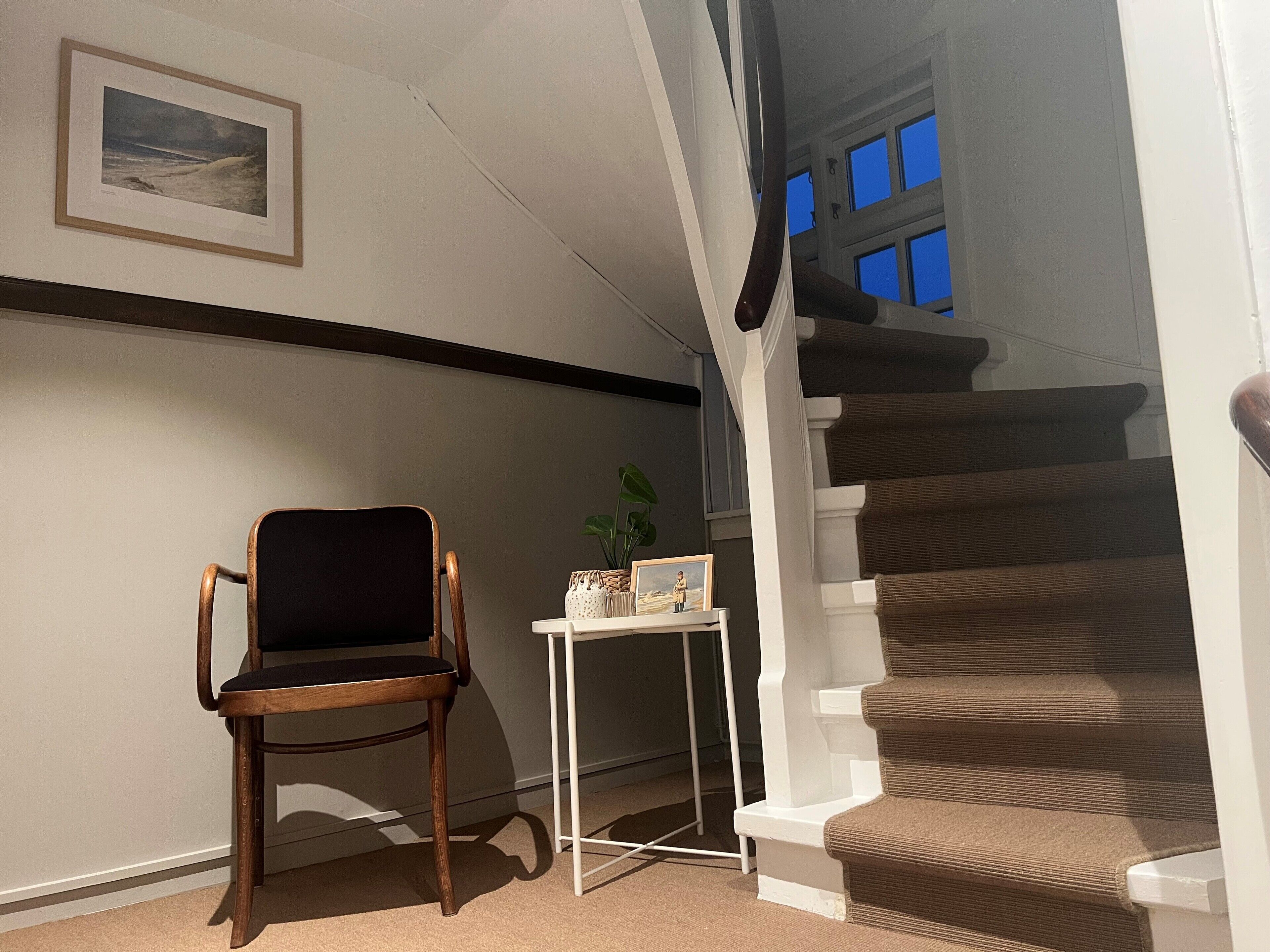 Una habitación con una silla de madera, una mesita con una planta e imagen, una escalera con escalones marrones y un cuadro en la pared.