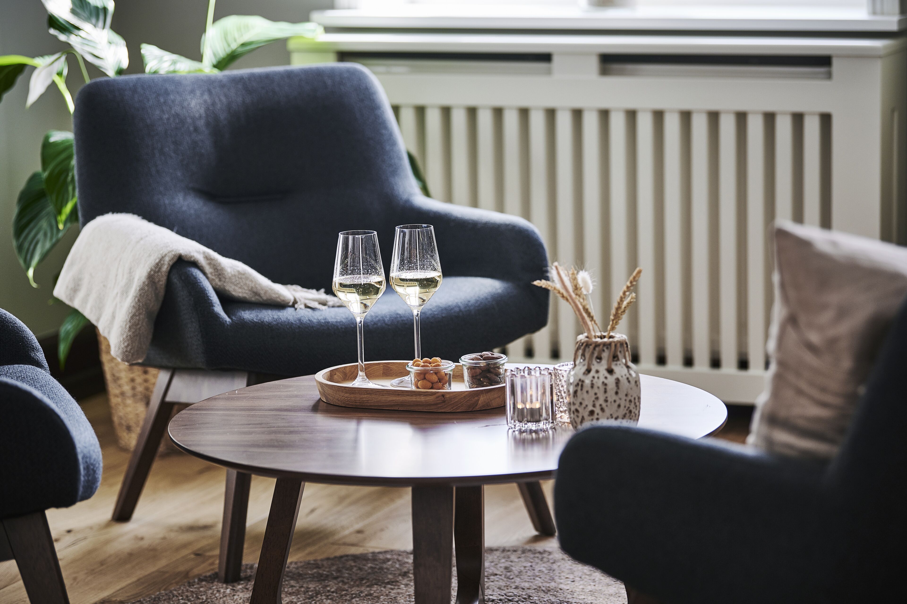 Un rincón acogedor con un sillón azul oscuro, una mesa redonda de madera con una bandeja de aperitivos y dos copas de vino blanco.