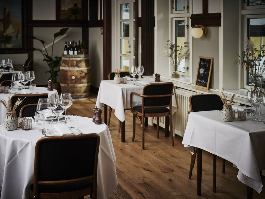 A restaurant with multiple tables set for dining, wooden flooring, and a view of the outdoors through large windows.